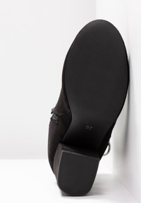 Semelle d'une botte noire en daim à la cheville avec talon bloc, taille 37 visible sur une semelle lisse, positionnée debout contre une surface blanche.