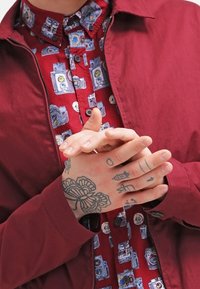 Burgundy zip-up jacket over a red shirt featuring a camera pattern. Hands with various black and blue tattoos positioned together.