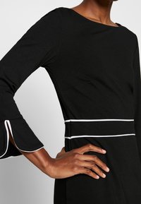 Schwarzes Kleid mit rundem Ausschnitt, langen Ärmeln mit weißem Besatz und tailliertem Design, das durch ein weißes Band betont wird. Glatter Stoff.