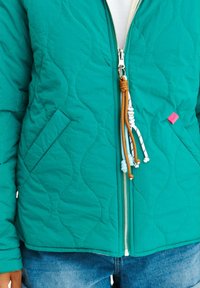 Tyrkysová prešívaná bunda so zipsom, ktorá má kontrastné šnúrky a malú ružovú akcentovú nálepku. Obsahuje bočné vrecká.