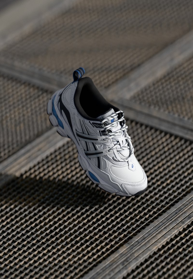Chaussure de sport blanche et bleue avec des accents noirs flottant au-dessus d'une surface en grille métallique texturée à la lumière naturelle.