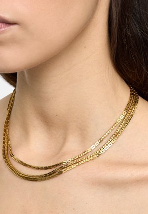 Zbliżenie na szyję i obojczyk osoby noszącej trzy warstwy złotych łańcuszków z prostokątnymi ogniwami.