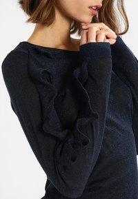 Granatowy sweter z falbaniastymi akcentami na rękawach, wykonany z materiału o teksturowanym splocie z delikatnymi, błyszczącymi nitkami.