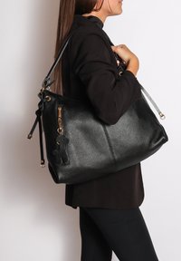Sac fourre-tout en cuir noir avec une finition texturée, doté de quincaillerie en or, d'une poche zippée et d'un charme décoratif accroché.
