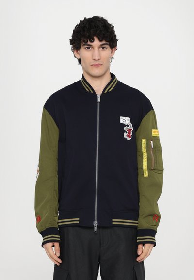 Bomberjacke mit einem marineblauen Körper und olivgrünen Ärmeln, ausgestattet mit Aufnähern, gestreiften Rippbündchen und einem Frontreißverschluss.