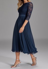 Kvinna i en marinblå spetsklänning med trekvartslånga ärmar och en svängig kjol, kombinerad med silverhöga klacksandaler med remmar.