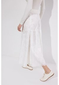 Γυναίκα με λευκό μακρυμάνικο τοπ, λευκή φούστα με δαντέλα, και λευκά κορδονιαστά παπούτσια, περπατά μπροστά σε λευκό τοίχο.