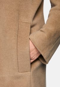 Beige Wollmantel mit weicher Textur, einer Seitentasche und einem taillierten Ärmel-Design mit glatter Oberfläche.