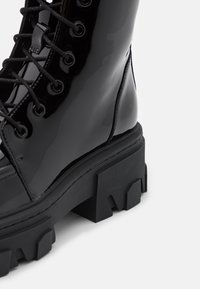 Botte de combat en cuir verni noir brillant avec semelle à crampons épaisses, détails cousus et lacets noirs sur fond blanc.