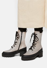 Bottines lacées grises et noires avec une semelle épaisse, fermeture éclair sur le côté et détail de surface texturé. Matériau durable avec des accents contrastés.