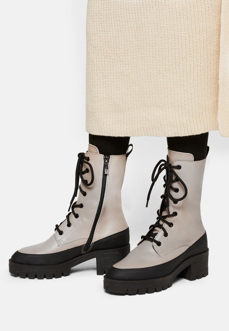 Bottines lacées grises et noires avec une semelle épaisse, fermeture éclair sur le côté et détail de surface texturé. Matériau durable avec des accents contrastés.