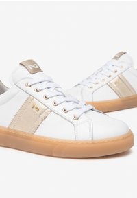 White sneakers with a textured finish, gold accents, and a transparent gum sole. Features white laces and branding on the tongue.