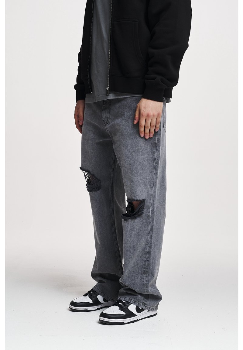 Jean gris usé avec des déchirures effilochées aux genoux, associé à un sweat à capuche noir zippé et des baskets noires et blanches, affichant une coupe décontractée.