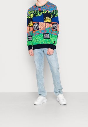 Uomo con maglione colorato a motivo con la scritta "Carlo Colucci", jeans azzurri e sneakers bianche, in piedi davanti a uno sfondo neutro.