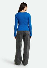 Top lungo a maniche lunghe blu a costine, design aderente, scollatura rotonda, abbinato a jeans grigi a vita alta con gamba larga, dettagli di cucitura visibili sui jeans.