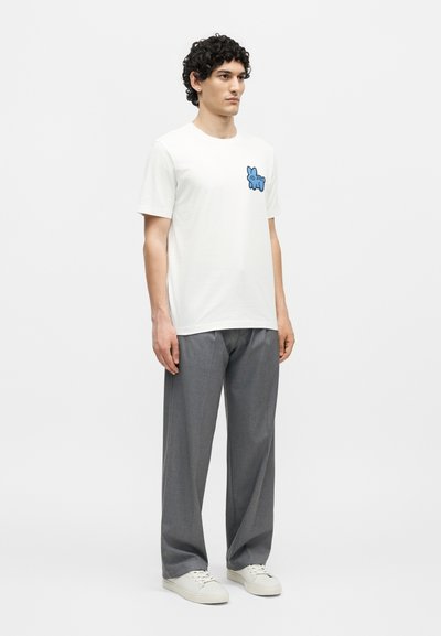 Άνδρας στέκεται φορώντας λευκό T-shirt με μικρό μπλε κινούμενο σκύλο στο στήθος, γκρι παντελόνια φαρδιάς γραμμής και λευκά αθλητικά παπούτσια σε απλό φόντο.