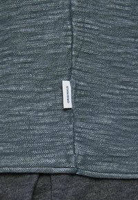 Gros plan sur un tissu bleu-gris texturé avec une couture verticale et une petite étiquette blanche portant la mention "ORIGINALS" attachée près du centre.
