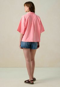 Loose-fit, short-sleeved pink shirt with a collar, paired with denim shorts and brown sandals. Smooth fabric with a casual cut.