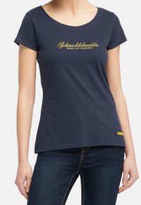 Laivansininen lyhythihainen t-paita, tehty puuvillasta, jossa on kultaiset tekstit "Schmuddelwedda" ja pieni keltainen tunnus helman kohdalla. Klassinen pyöreä kaula-aukko.