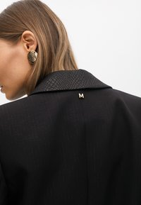 Kobieta z włosami sięgającymi ramion w kolorze brązowym, nosząca czarną, fakturowaną marynarkę oraz złotą broszkę w kształcie litery "M" i dużymi, fakturowanymi złotymi kolczykami.