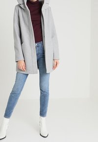 Osoba w jasnoszarej kurtce z kapturem, burgundowym golfie, niebieskich dżinsach i białych botkach za kostkę, stojąca na tle jednolitego jasnego tła.