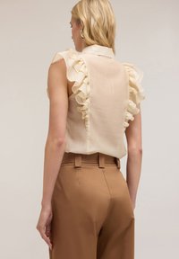 Beige ärmlös blus med volangdetaljer på axlarna, mjukt tyg och krage, matchad med bruna höga byxor.