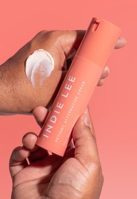 Persikanvärinen pumppullomerkki "INDIE LEE RETINOL ALTERNATIVE CREAM", pidetään käden yllä, johon on levitetty kermamaista tuotetta.