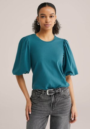 Kihara juustega naine, kes kannab sinakashalli lukuga varrukaga pluusi, musta naelutatud vööd ja tumedat halli teksapükse ühevärvilise tausta ees.