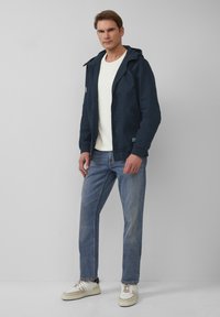Marineblau gefütterte Jacke mit Frontreißverschluss, zwei Taschen an den Seiten und elastischen Bündchen, kombiniert mit einem leichten Pullover, blauen Jeans und weißen Sneakers.