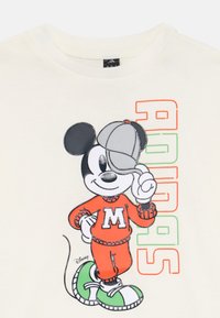 Mickey Mouse con un traje naranja y un logo de 'M', inclinando un sombrero gris, al lado de un texto colorido de "ADIDAS" sobre un fondo crema.