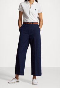 Vit polo skjorta med en marinblå emblem, marinblå vida byxor, brun vävd bälte och vita sneakers med en färgad rand.