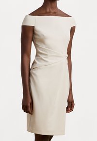 Cremefärgad off-the-shoulder klänning med figurnära överdel, tvinnad detalj vid midjan och knä-lång nederkant. Slät och elegant tygtextur.
