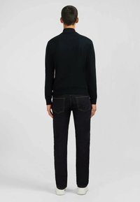 Pull en tricot noir avec un col montant, associé à un jean bleu foncé et des baskets blanches. La vue arrière montre une texture lisse et un design ajusté.