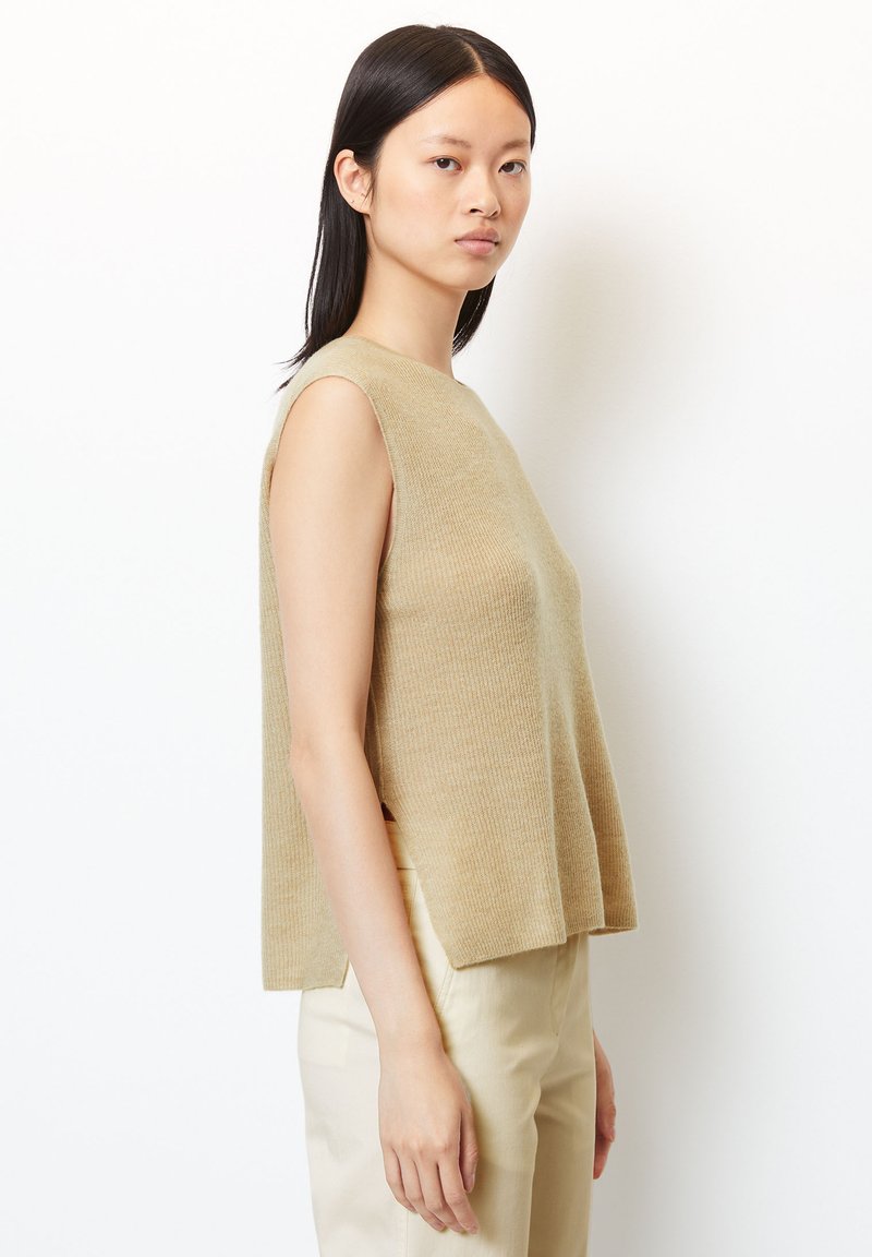 Junge Frau mit glattem schwarzen Haar, die ein ärmelloses beiges Strickoberteil und helle beige Hosen trägt, vor einem weißen Hintergrund steht.