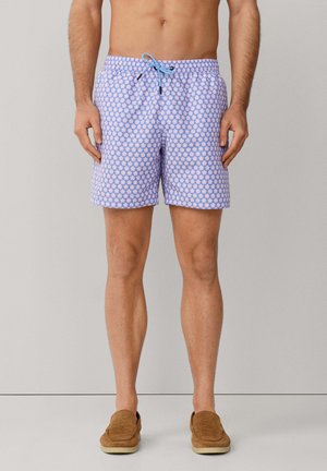 Ein Mann trägt blaue und pink gemusterte Badeshorts mit Kordelzug und steht barfuß vor einem schlichten Hintergrund.