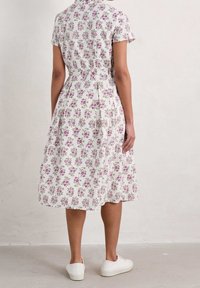 Vit blommig klänning med korta ärmar, en figursydd midja och en urflackad kjol. Dekorerad med små rosa blommor och gröna blad. Matchad med vita skor.