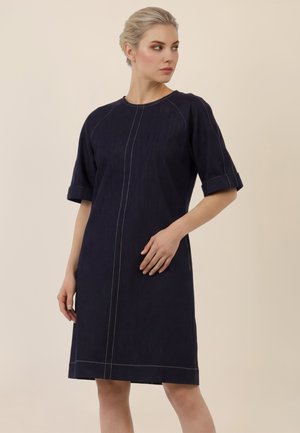 Vrouw staand, gekleed in een marineblauwe jurk tot op de knie met halve mouwen en witte verticale en horizontale stikdetails.