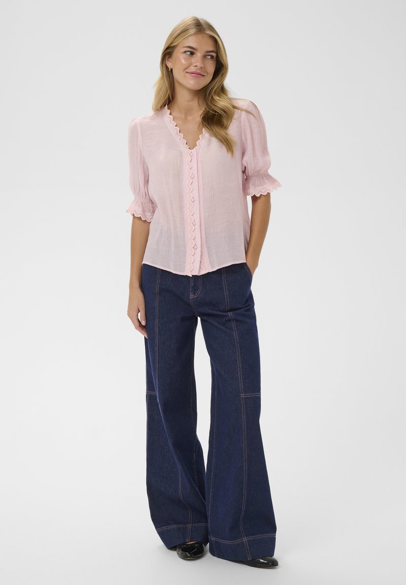 Blouse rose clair à bords festonnés et manches bouffantes, associée à un pantalon en jean large et sombre, souligné par des coutures contrastées.