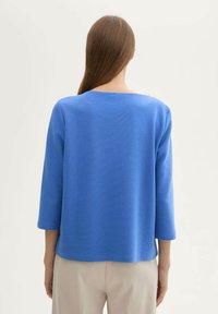 Blaues, strukturiertes Langarmtop mit einem Bootsausschnitt, lockerer Passform und überschnittenen Schultern, kombiniert mit hellen Hosen.