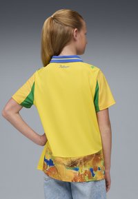 Camisa polo amarilla con acentos verdes y azules, que presenta un patrón texturizado y colorido en el dobladillo y un logo de la marca bordado en el cuello.