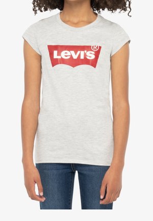 Dziewczyna w jasnoszarej koszulce Levi’s z czerwonym logo i niebieskich dżinsach, stoi z rękoma swobodnie opuszczonymi wzdłuż ciała.