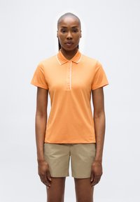 Femme debout, portant un polo à manches courtes orange avec des boutons et un short beige, sur un fond clair uni.