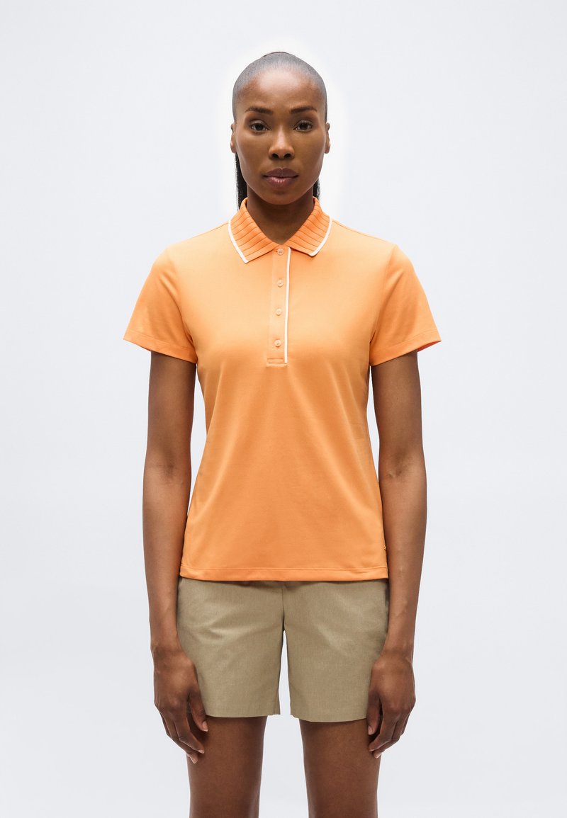 Femme debout, portant un polo à manches courtes orange avec des boutons et un short beige, sur un fond clair uni.