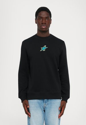 Man wearing a black sweatshirt with a small green and blue star emblem on the chest, paired with light blue jeans, standing against white background.