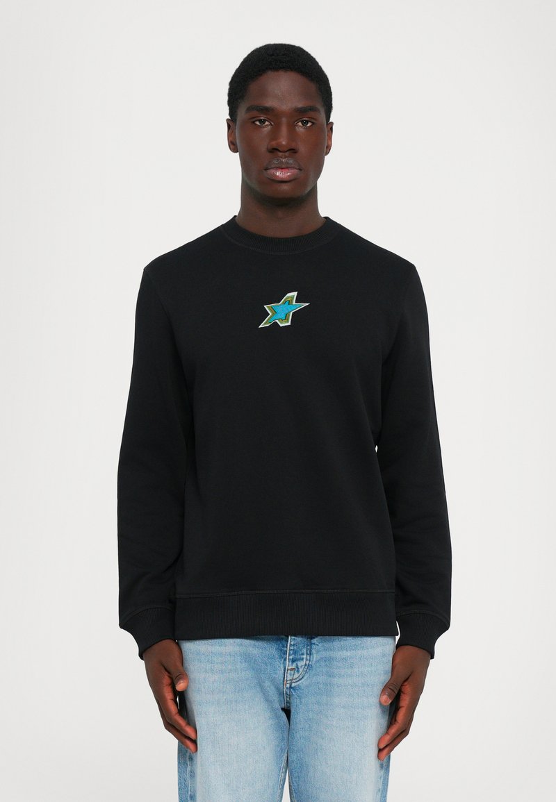 Man wearing a black sweatshirt with a small green and blue star emblem on the chest, paired with light blue jeans, standing against white background.