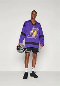 Vyras, apsirengęs purpurine Los Andželo "Lakers" marškinėliais ir juodais krepšinio šortais, laiko juoda-balta krepšinio kamuolį prieš vienodą foną.