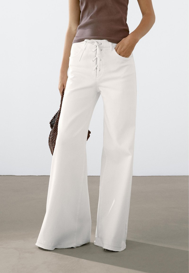 Witte hoge taille brede broek met een veter sluiting aan de voorkant, twee zijzakken en een gladde textuur, gecombineerd met een bruine top en een gehaakte tas.