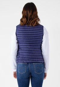 Gilet imbottito blu navy con design trapuntato, caratterizzato da cuciture orizzontali, colletto alto e vestibilità slim, indossato sopra una camicia bianca a maniche lunghe.