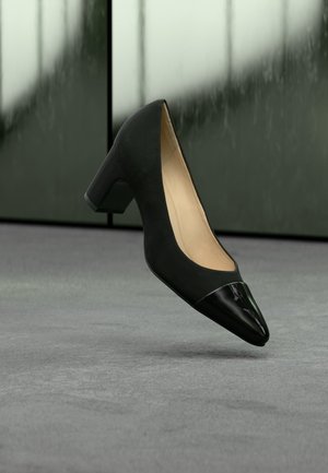 Czarny damski but na średnim obcasie z połyskującym, spiczastym noskiem, unoszący się nad szarą podłogą na tle rozmytego ciemnozielonego tła.