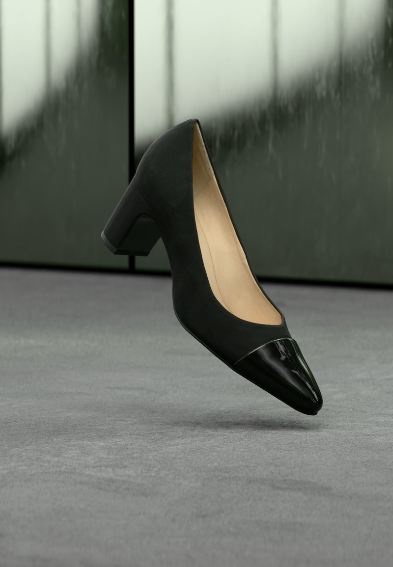 Μαύρο γυναικείο παπούτσι με μεσαίο τακούνι και γυαλιστερή μυτερή μύτη, να αιωρείται πάνω από γκρι πάτωμα με φόντο θολό σκούρο πράσινο.